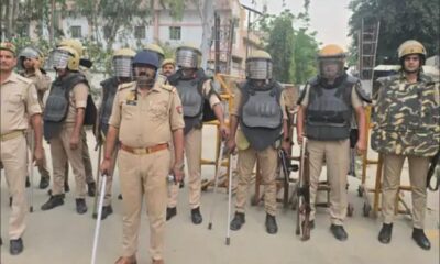 Mandi Dhanaura mein police ki tukdi riot-gear aur lathis ke saath, pichhe white residential building.