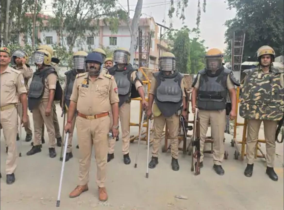 Mandi Dhanaura mein police ki tukdi riot-gear aur lathis ke saath, pichhe white residential building.
